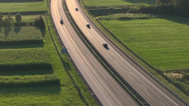 AERIAL: A flying shot of golden sun shining upon a highway, surrounded by lush green fields. Matured crops of corn seat readily on the fields. A few sunlit cars pass by peacefully on their commute.