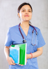 Positive confident young woman, professional doctor, in blue uniform and with phonendoscope around neck holding medical records. Studio photoshoot on gray background - obrazy, fototapety, plakaty