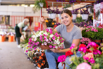 Cheerful young girl choosing ornamental plants to decorate courtyard at flower market, looking with interest at flowering petunias in hanging cache-pots - 769229855