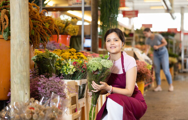 Portrait of a positive female flower market seller with a bouquet of daisies for buyers - 769230409