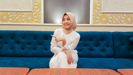 young asian woman candid smile expression hand pointing to right sitting on sofa with islamic...