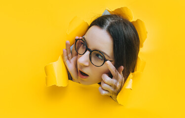 Portrait of a young woman with glasses looking through a hole in yellow paper. An incredulous look....