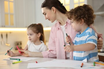 Mother and her little children drawing with colorful markers at table in kitchen