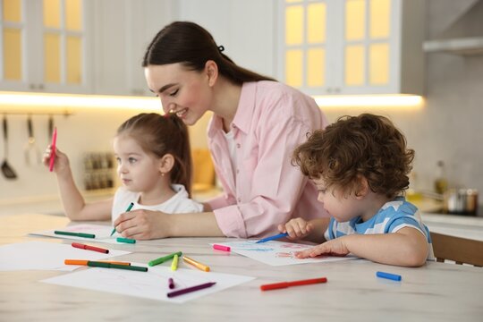 Mother and her little children drawing with colorful markers at table in kitchen