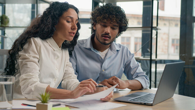 Arabian female leader coach help intern explain paperwork and financial data business strategy consult client. Smiling Indian colleague businessman talk with businesswoman corporate teamwork at office