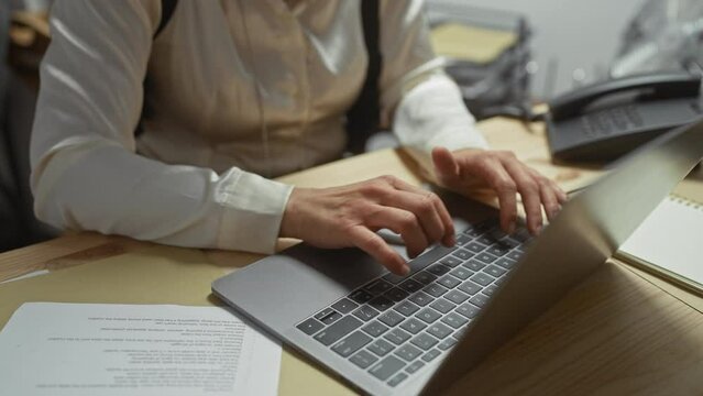 A mature woman in an office types on a laptop with documents and a phone visible, suggesting business or administrative work.
