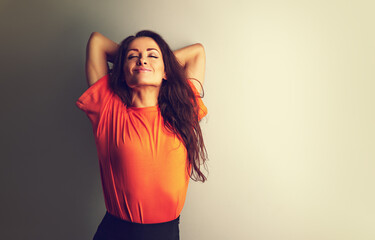 Beautiful young relaxing dreaming smiling woman in casual clothing with closed eyes with arms behind head in orange casual shirt on grey studio background on empty copy space. Closeup