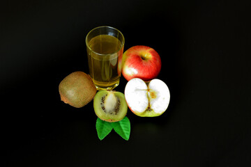 A glass of fresh fruit juice on a black background, next to a ripe red apple and pieces of kiwi fruit.