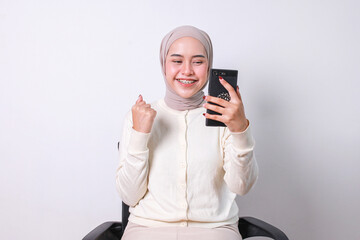 Young woman in hijab looking at smartphone and clenching fist showing victory gesture hand