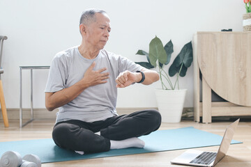 Senior Asian man sitting on floor and looking at watch after exercise at home