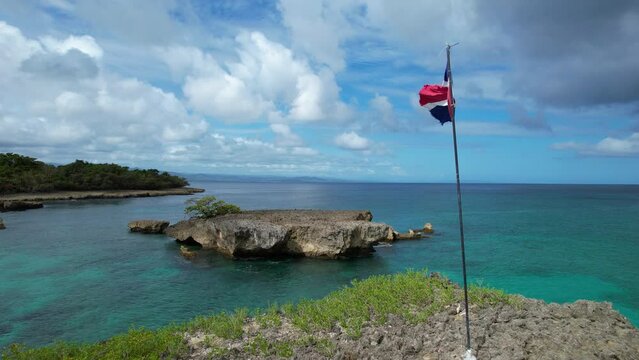 Aerial approaching shot of flag of Dominican Republic on boulder and Caribbean sea in Background