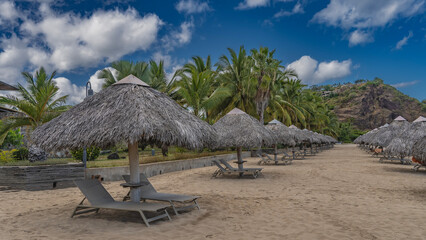 Sun loungers stand in rows under straw sun umbrellas on the sandy beach. Palm trees and hills against a background of blue sky and clouds. Madagascar. Nosy Be