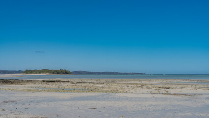 The low tide. The sand and rocks of the seabed were exposed. Turquoise ocean, mountains in the distance. Tiny silhouettes of people walking on the beach are visible. Clear blue sky. Copy space.