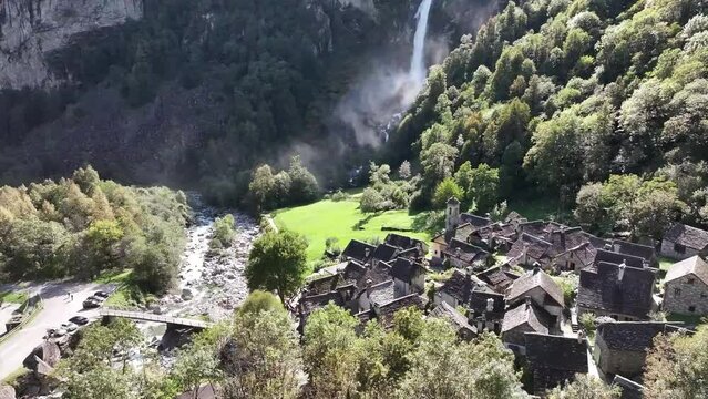 An aerial captures the majestic Foroglio Waterfall in Maggiatal, Tessin, Schweiz. Sunlight illuminates the cascading water against the backdrop of a quaint ancient village nestled in a valley below.