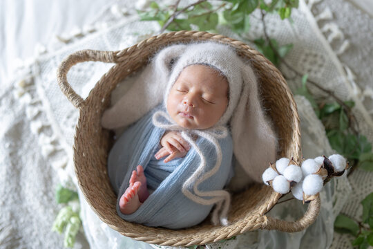 Newborn photo of a newborn baby who is 8 days old, half Taiwanese and half Australian, wrapped in a blue wrap, wearing a rabbit hat and sleeping in a basket. 生後8日の台湾人とオーストラリア人のハーフの新生児の赤ちゃんが青いおくるみを巻かれて