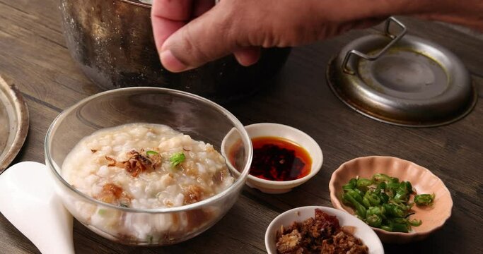 Asian thick rice porridge bubur chicken beef meat fish prawn rustic silver pot ladle serving in glass bowl fried shallots green onion chilli oil condiment