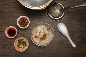 Asian thick rice porridge bubur chicken beef meat fish prawn rustic silver pot ladle serving in glass bowl fried shallots green onion chilli oil condiment