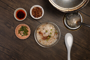 Asian thick rice porridge bubur chicken beef meat fish prawn rustic silver pot ladle serving in glass bowl fried shallots green onion chilli oil condiment