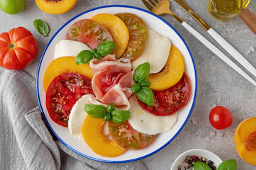 Salad of sliced multi-colored tomatoes, fresh peaches, mozzarella, prosciutto and basil on a gray concrete background. Delicious summer salad.