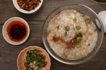 Asian thick rice porridge bubur chicken beef meat fish prawn rustic silver pot ladle serving in glass bowl fried shallots green onion chilli oil condiment