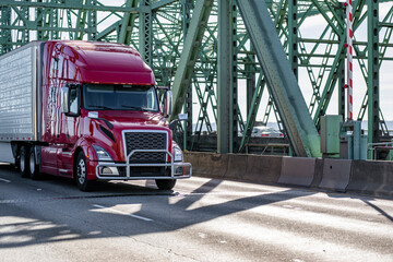 Dark red big rig bonnet semi truck transporting cargo in refrigerator semi trailer driving on the arched truss bridge
