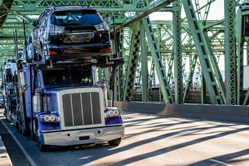 Heavy duty big rig blue car hauler semi truck transporting cars on two level modular semi trailer driving on the truss bridge at sunny day