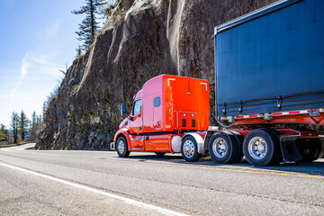 Industrial red big rig shiny semi truck transporting cargo in covered dry van semi trailer running on the mountain winding road with rock cliff