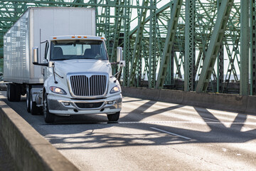 Local carrier white day cab big rig semi truck transporting cargo in dry van semi trailer driving on the arched truss bridge at sunny day