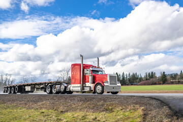 Powerful classic red big rig semi truck tractor with chrome parts transporting empty flat bed semi trailer driving on the wet after the rain narrow road