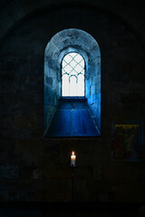 Fototapeta premium A single small, burning candle stands in front of a church window with a mosaic, light shining through the window (3787)