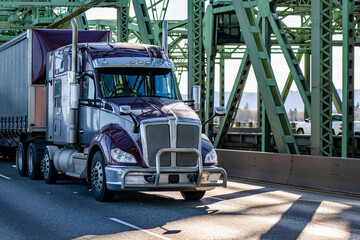 Purple big rig bonnet semi truck with grey accents transporting cargo in dry van tarp semi trailer running on the truss arched bridge across the Columbia River