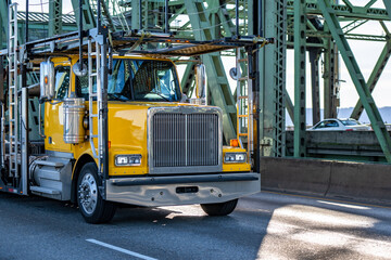 Yellow classic bonnet big rig semi truck tractor with empty two level hydraulic semi trailer driving on the truss arched bridge