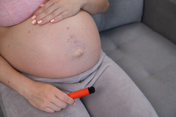 A pregnant woman smokes a vape while sitting on the couch. Close-up of the belly with allergies.