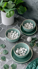 Obraz na płótnie Canvas Three bowls of white and blue ceramic are on a table with a green tablecloth. The bowls are filled with small white objects, and there is a potted plant in the background. The scene has a calm