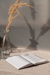 Open book and vase with dried grass on table with beige linen tablecloth, summer boho still life,...