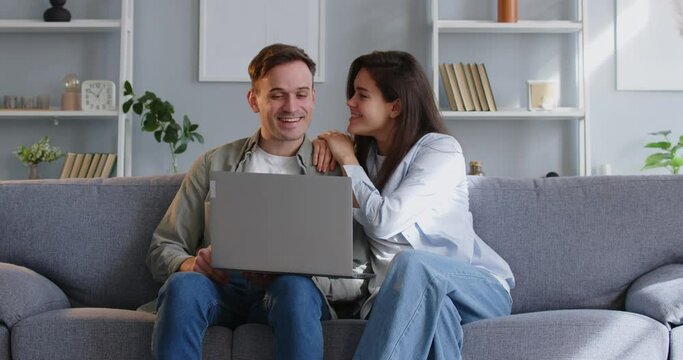 Happy young family couple using a laptop computer. Cheerful wife sits down on the sofa next to her husband who is online shopping on his notebook PC and they continue together