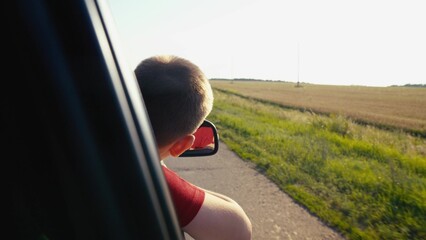 child boy son enjoys summer day with his hand out car window, car rushing along road, child feels...