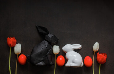 Dark Eastern photo with two bunnies black and white, red eggs and red and white flowers on black...