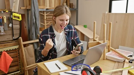 A young caucasian blonde woman celebrates success on her smartphone in a woodworking workshop with tools and a laptop around.