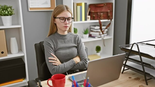 A pensive young woman with blonde hair and glasses crosses her arms while working in a modern office