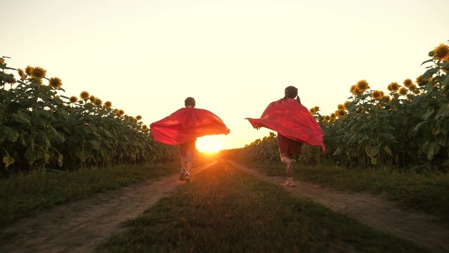 Boy and girl superhero in red cloak running flying on road at sunflower field sunset back view. Happy kids playing fantasy imagination pretend powerful hero character planet protect enjoy childhood