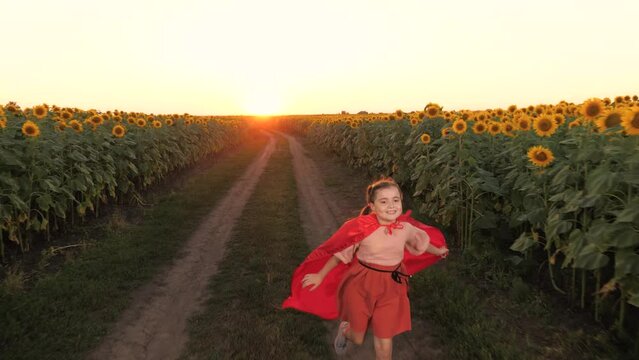 Smiling girl in red cloak superhero costume image flying at sunflower field sunset plantation. Happy female teen kid child hero running flight imagination fantasy at agriculture harvest flower meadow
