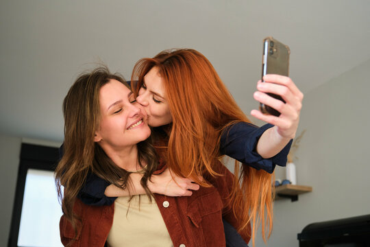 Lesbian couple kissing and taking a selfie, piggyback at home.