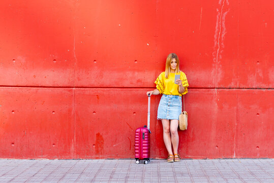 young traveler stands against a red wall, her suitcase at her side