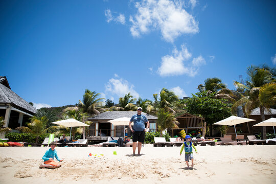 Dad in front of Caribbean Beach Bar Watches Young Sons Play in Sand