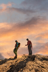 Two men are standing on a rocky mountain, one of them holding a rope. The sky is orange and the clouds are low, creating a moody atmosphere
