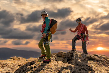 Two people are on a mountain, one of them is holding a rope. The sun is setting in the background, creating a warm and peaceful atmosphere - Powered by Adobe
