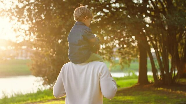Dad and Son Walking in a Park at Sunset.
The Son is Sitting on the Father's Shoulders, They Are Having Fun and Playing Together