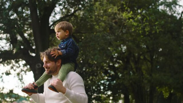 Dad and Son Walking in a Park at Sunset.
The Son is Sitting on the Father's Shoulders, They Are Having Fun and Playing Together