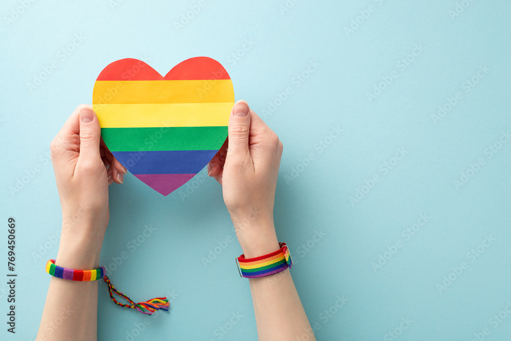 Wall mural Rainbow heart-shaped card held by a woman's hands wearing symbolic bracelet and wristlet in a first-person top view on a pastel blue background with space for text or advertising